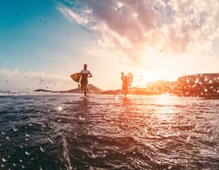 Happy Urfers Running With Surf Boards On The Beach - Sporty People Having Fun In Sunny Day - Extreme Sport, Travel And Vacation Concept - Focus On Bodies Silhouettes - Water On Camera Lens Wall Mural