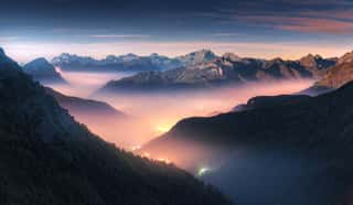 Mountains In Fog At Beautiful Night In Autumn In Dolomites, Italy  Landscape With Alpine Mountain Valley, Low Clouds, Forest, Colorful Sky With Stars, City Illumination At Dusk  Aerial  Passo Giau Wall Mural