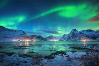 Aurora Borealis Over The Sea Coast, Snowy Mountains And City Lights At Night  Northern Lights In Lofoten Islands, Norway  Starry Sky With Polar Lights  Winter Landscape With Aurora Reflected In Water Wall Mural